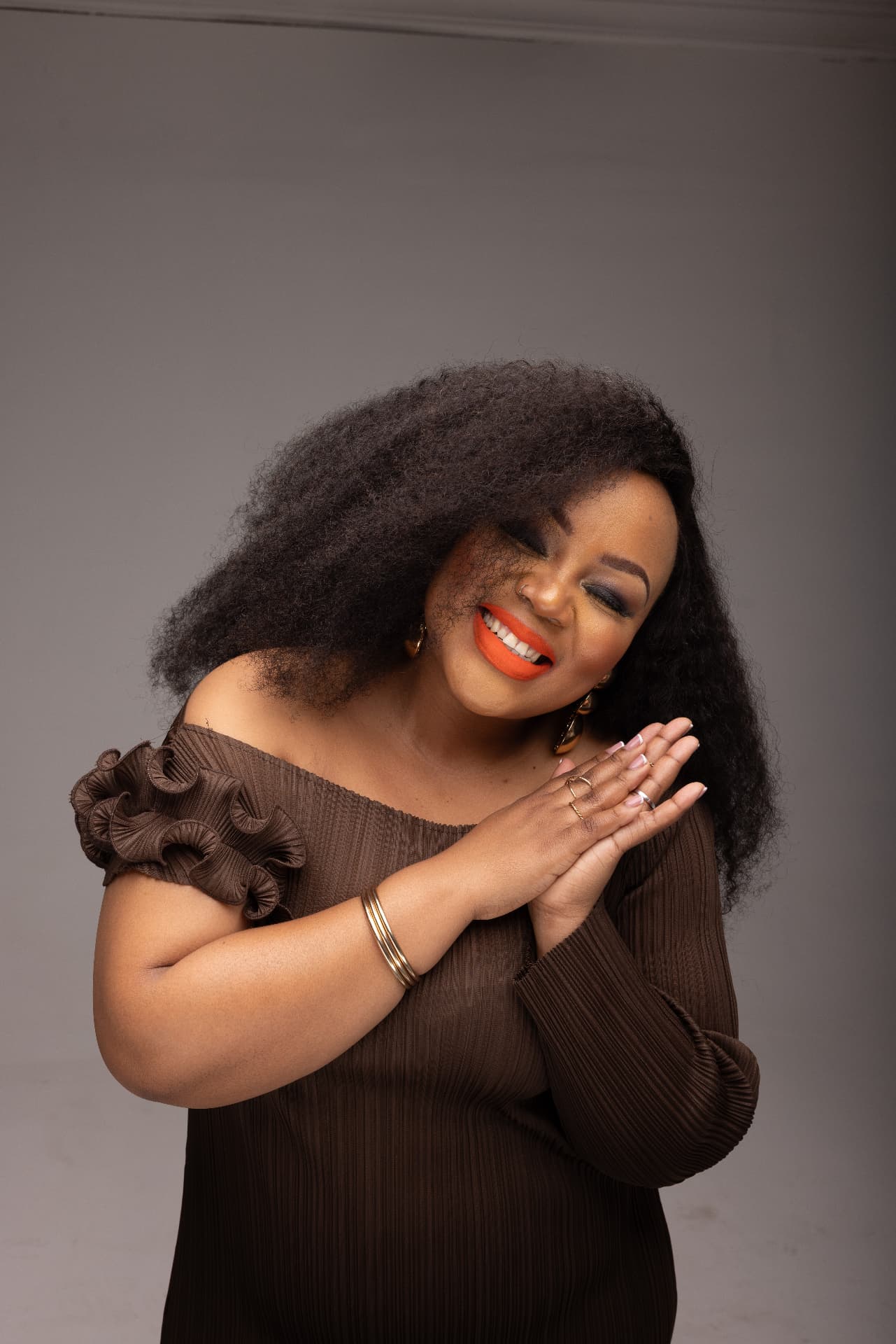 Lebo Thabethe smiling with hands clasped in a brown ruffled dress